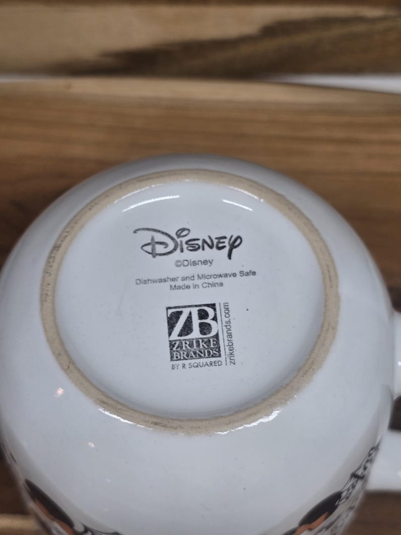 May include: White ceramic Disney mug with a black and white graphic. The bottom of the mug features the Disney logo, along with text indicating it is dishwasher and microwave safe, and the Zrike Brands logo. Made in China.
