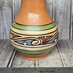 May include: A terracotta vase with a flared rim and a rounded body featuring horizontal bands of green, cream, and a black-outlined pattern with blue and brown accents. A Westcott ruler is positioned at the base, showing a width of 6 inches.