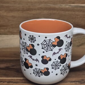 May include: White ceramic mug with an orange interior, decorated with black Mickey Mouse silhouettes with orange bows, spiderwebs, bats, and stars. The mug has a white handle and is perfect for Halloween.