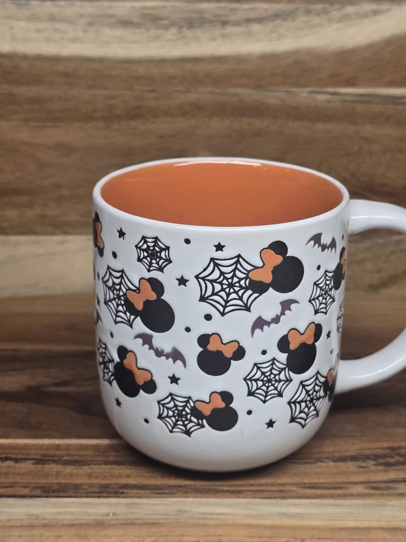 May include: White ceramic mug with an orange interior, decorated with black Mickey Mouse silhouettes with orange bows, spiderwebs, bats, and stars. The mug has a white handle and is perfect for Halloween.