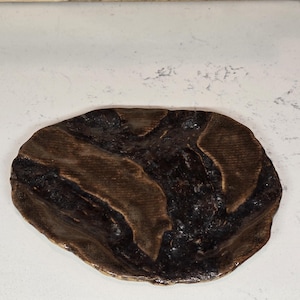 May include: A handmade, organic-shaped ceramic plate with a dark brown and black mottled glaze. The plate has a textured surface and a slightly raised rim, perfect for serving appetizers or small dishes.