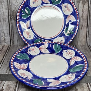 May include: Two round ceramic plates featuring a white center and a wide blue rim decorated with hand-painted white fish with pink fins and green seaweed accents. The outer edge of each plate is finished with a repeating pattern of small red dots.