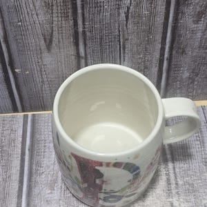 May include: White ceramic mug with a rounded body and a handle. The mug features a colorful, festive design with red, blue, and green accents. The interior of the mug is a clean, bright white.