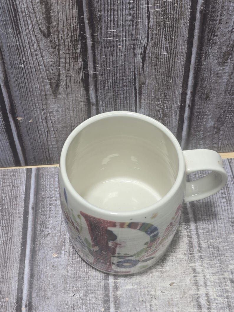 May include: White ceramic mug with a rounded body and a handle. The mug features a colorful, festive design with red, blue, and green accents. The interior of the mug is a clean, bright white.