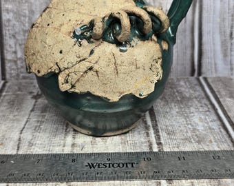 May include: A dark green ceramic pitcher with a glossy finish, featuring a textured, light tan leaf-shaped appliqué on the front. The pitcher has a curved handle and a narrow neck. A metal ruler shows the base width is approximately 6 inches.