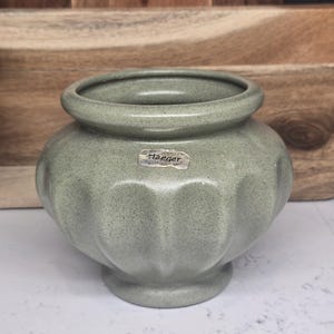 May include: A sage green ceramic vase with a textured surface and a rounded shape. The vase has a wide opening and a decorative, fluted design around the body. A small label is affixed to the front. The vase is a decorative home decor item.