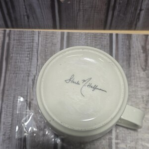 May include: A white ceramic mug with a handle, viewed from the bottom. The base of the mug features a handwritten signature in black ink, "Starla M. Halfmann". The mug is set against a backdrop of weathered wooden planks.