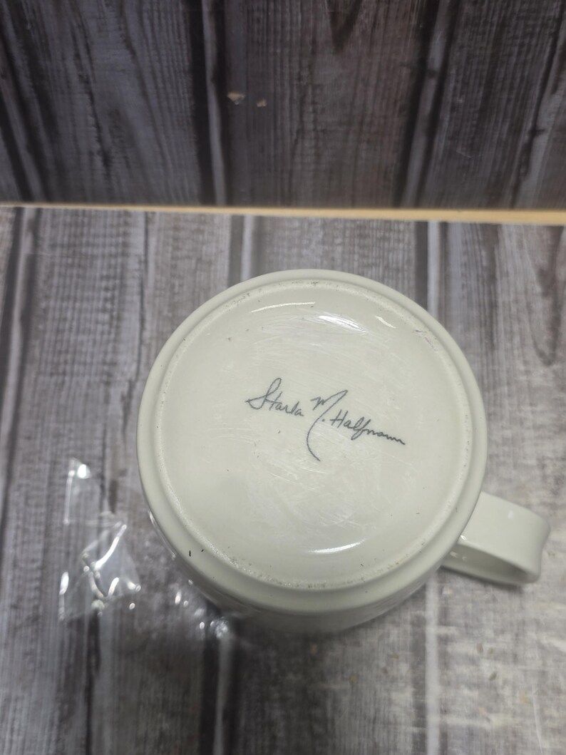 May include: A white ceramic mug with a handle, viewed from the bottom. The base of the mug features a handwritten signature in black ink, "Starla M. Halfmann". The mug is set against a backdrop of weathered wooden planks.