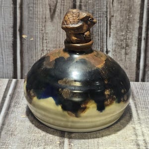 May include: A ceramic bottle with a turtle-shaped stopper. The bottle has a round shape with a black and brown glazed top, transitioning to a cream-colored base. The turtle stopper is brown and detailed.