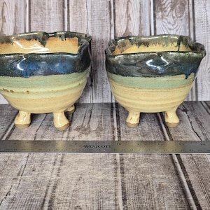 May include: Two ceramic bowls with three small feet each, featuring a cream-colored base, a light green middle band, and a dark blue glazed rim. A metal Westcott ruler is placed in front of the bowls, showing a scale in inches and centimeters.