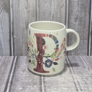 May include: Cream-colored ceramic mug with a large, colorful letter "P" on the front. The letter is decorated with a floral design and surrounded by colorful flowers and leaves. The mug has a curved shape and a handle.