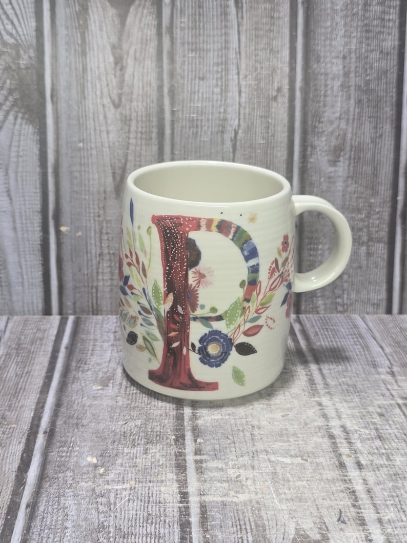 May include: Cream-colored ceramic mug with a large, colorful letter "P" on the front. The letter is decorated with a floral design and surrounded by colorful flowers and leaves. The mug has a curved shape and a handle.