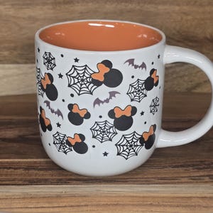 May include: White ceramic mug with a Halloween theme. The mug features a black and orange design of Mickey Mouse-inspired silhouettes with spiderwebs, bats, and stars. The interior of the mug is orange, and it has a white handle.