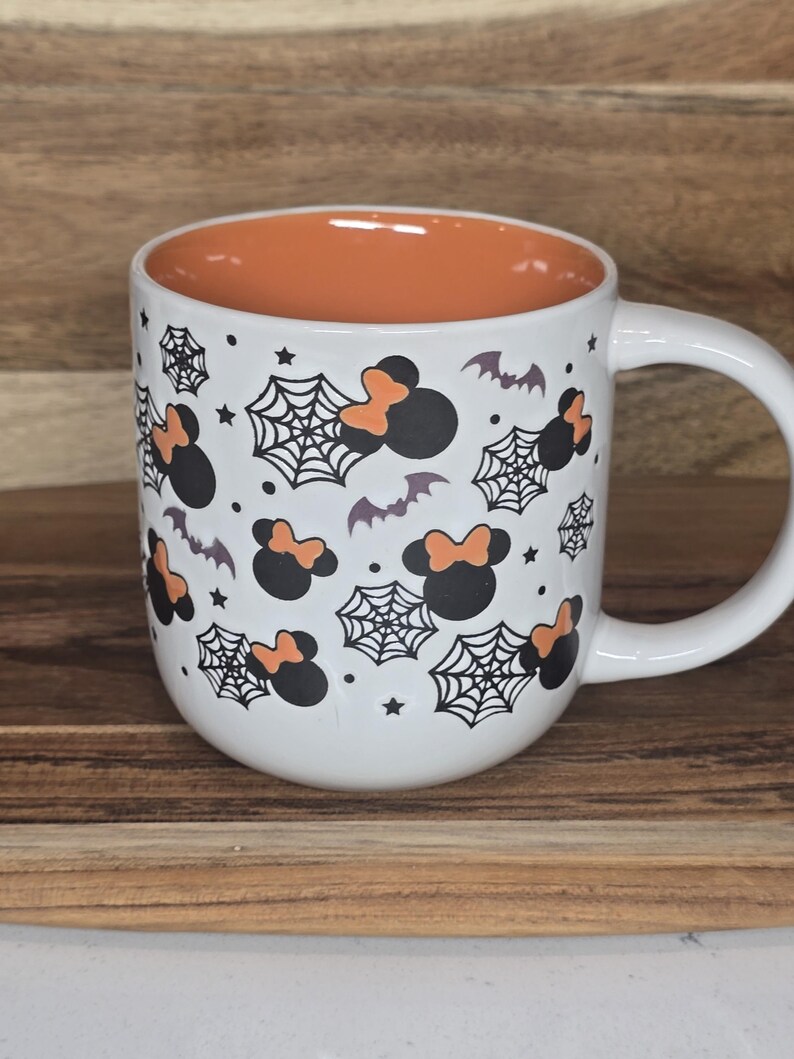 May include: White ceramic mug with a Halloween theme. The mug features a black and orange design of Mickey Mouse-inspired silhouettes with spiderwebs, bats, and stars. The interior of the mug is orange, and it has a white handle.