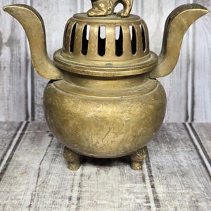 May include: A brass incense burner with two curved handles and a lid topped with a small lion figure. The burner features a rounded body supported by three feet. A metal ruler placed in front shows the burner is approximately 6 inches wide.