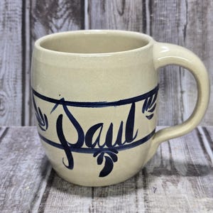 May include: Cream-colored ceramic mug with a rounded body and a large handle. The mug features the name "Paul" in dark blue script, encircled by a blue band with leaf-like designs. The mug is set against a wooden background.