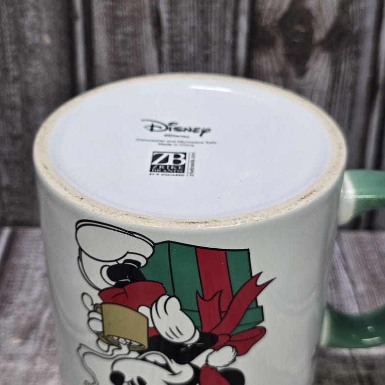 K&ouml;nnte beinhalten: Wei&szlig;e Disney-Keramiktasse mit Mickey Mouse, der ein verpacktes Geschenk mit roter Schleife h&auml;lt. Die Tasse hat einen gr&uuml;nen Griff, und das Disney-Logo ist unten sichtbar.