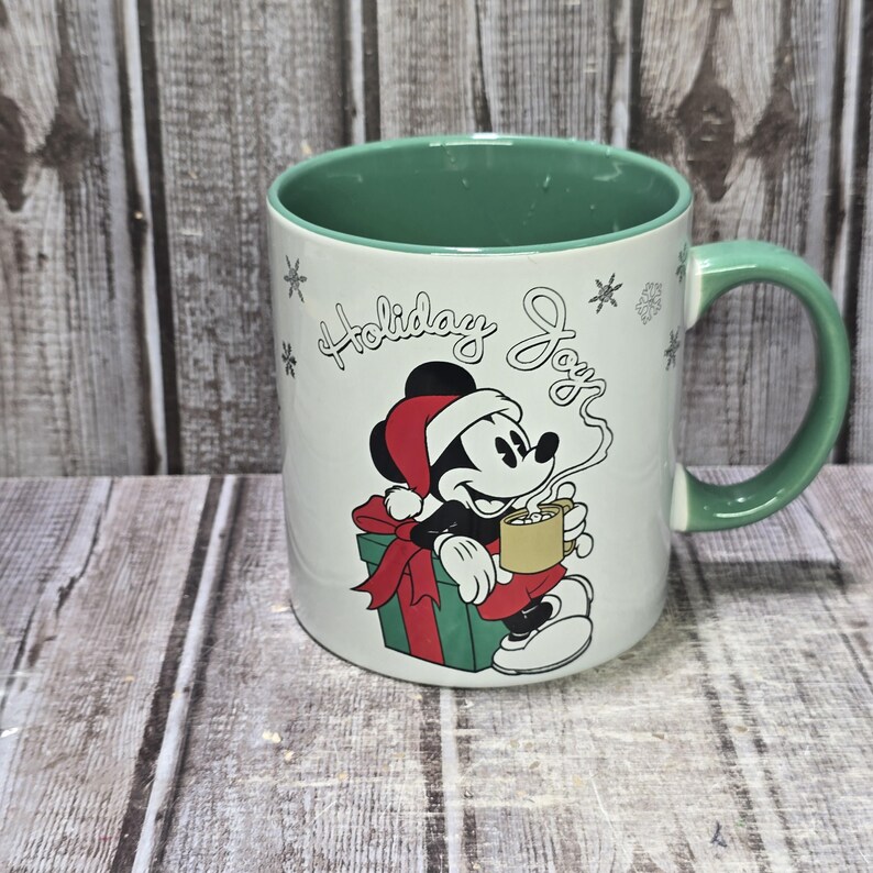 K&ouml;nnte beinhalten: Wei&szlig;er Keramikbecher mit gr&uuml;nem Innenraum und Griff. Der Becher zeigt eine Grafik von Mickey Mouse mit Weihnachtsm&uuml;tze, die eine Tasse Kakao h&auml;lt, mit den Worten "Holiday Joy" und Schneeflocken-Designs. Ein festlicher Becher zum Thema Feiertage.