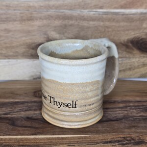 May include: A handmade ceramic mug with a beige and cream color scheme. The mug features the text "Know Thyself" and "II Ch. 33:12". The mug has a curved handle and is set against a wooden background.
