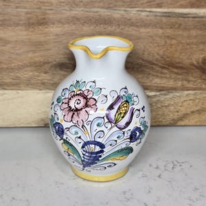 May include: A small, white ceramic vase with a yellow rim and base. The vase is adorned with a hand-painted floral design in shades of pink, purple, blue, and green. The vase is approximately 5 inches tall.