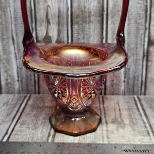 May include: A red iridescent carnival glass basket with a tall arched handle and a flared rim. The body features a pressed geometric starburst pattern and a hexagonal base. A metal ruler is positioned in front, showing a width of 6 inches.