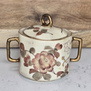 May include: Cream-colored ceramic sugar bowl with a lid, featuring a floral design in shades of pink, brown, and gray. The bowl has gold-colored handles and a gold-colored handle on the lid. The sugar bowl is a vintage item.