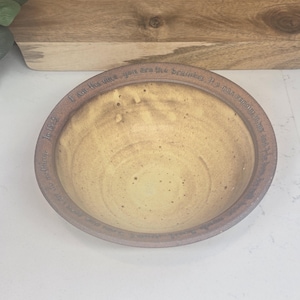May include: A handmade ceramic bowl with a light brown interior and a darker brown rim. The rim features an engraved inscription. The bowl is set against a neutral background, highlighting its earthy tones and craftsmanship.
