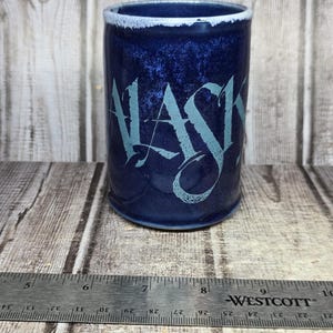 May include: A cylindrical ceramic mug with a deep blue mottled glaze and the word ALASKA printed in a light blue stylized font. A metal Westcott ruler is positioned in front of the mug, showing a width of approximately 3.5 inches.