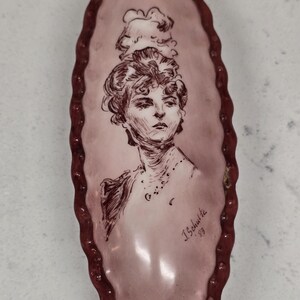 May include: An oval-shaped, pink ceramic dish with a portrait of a woman in a bonnet. The portrait is in brown ink, and the dish has a dark red, textured border. The artist's signature, "J. Schulte '83", is visible.