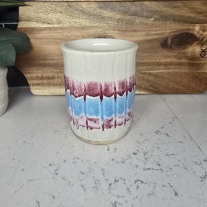 May include: A white ceramic utensil holder with a blue and burgundy striped design. The holder is sitting on a light gray countertop.