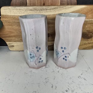 May include: Two pink ceramic mugs with a white and blue floral design. The mugs have a textured, wavy surface.