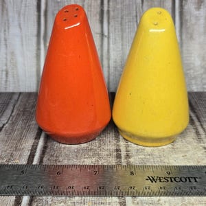 May include: A pair of cone-shaped salt and pepper shakers in bright orange and yellow ceramic, positioned behind a metal Westcott ruler that shows the set spans approximately 4 inches in width, with three small holes on top of each shaker.