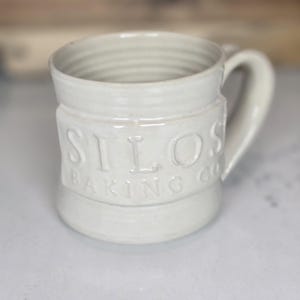 May include: Cream-colored ceramic mug with a curved handle. The mug features the embossed text "SILOS BAKING CO" around the middle. The mug has a textured, ridged interior and a smooth, glossy exterior. The mug is designed for beverages.
