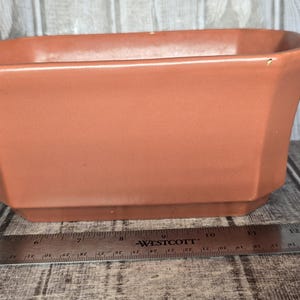 May include: A terracotta-colored ceramic planter with a hexagonal shape and a smooth finish. A metal Westcott ruler is positioned in front of the planter, showing the base width is approximately 7.5 inches and the top width is 9 inches.