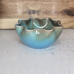 May include: A ceramic bowl with a ruffled rim, showcasing a gradient of colors. The bowl transitions from a light blue base to a turquoise and brown rim. The handmade bowl is likely used for serving or display.