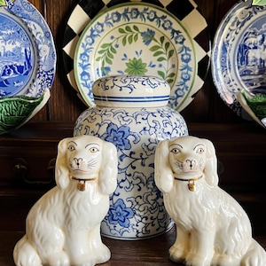 May include: Two white ceramic dog figurines with gold collars sit in front of a blue and white floral jar. Decorative plates with blue and green patterns are in the background, creating a classic, elegant display.