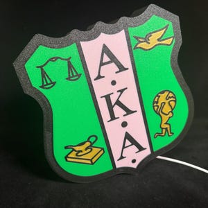 May include: A green, pink, and gold shield-shaped sign with the letters "A.K.A." in the center. The shield is divided into three sections, each with a different symbol: a scale, a lamp, and a globe.