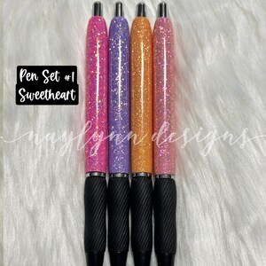 May include: Set of four glitter pens with black grips. The pens are pink, purple, orange, and pink. The text "Pen Set #1 Sweetheart" is visible in the image.