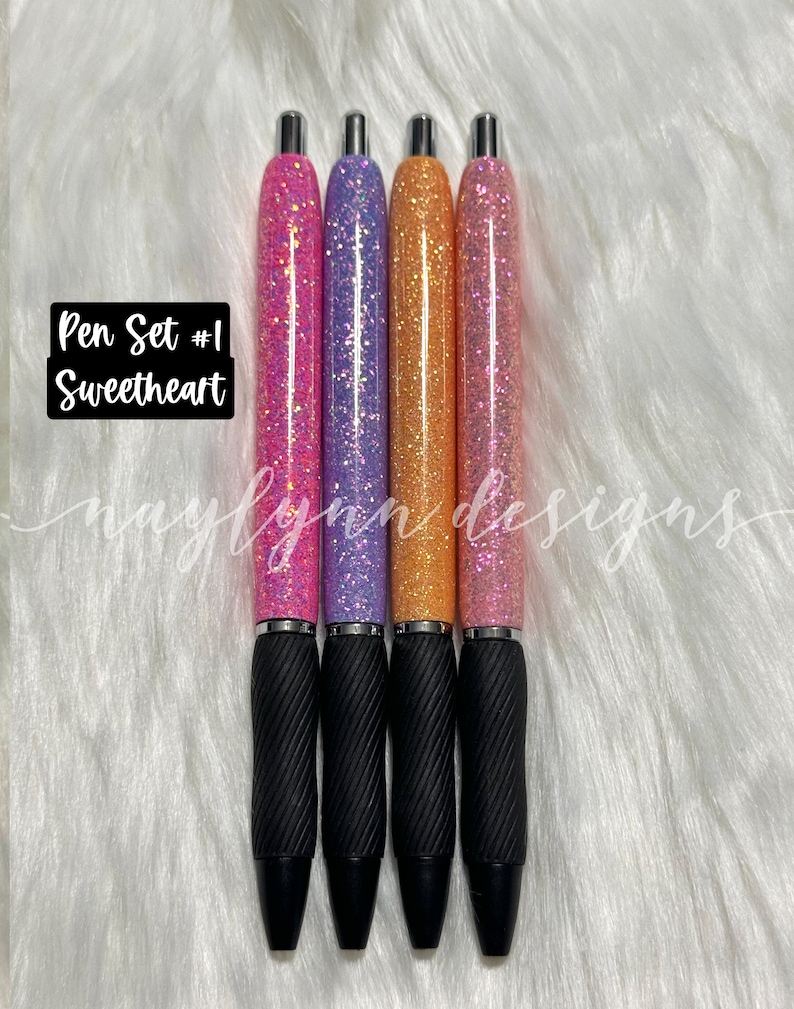May include: Set of four glitter pens with black grips. The pens are pink, purple, orange, and pink. The text "Pen Set #1 Sweetheart" is visible in the image.