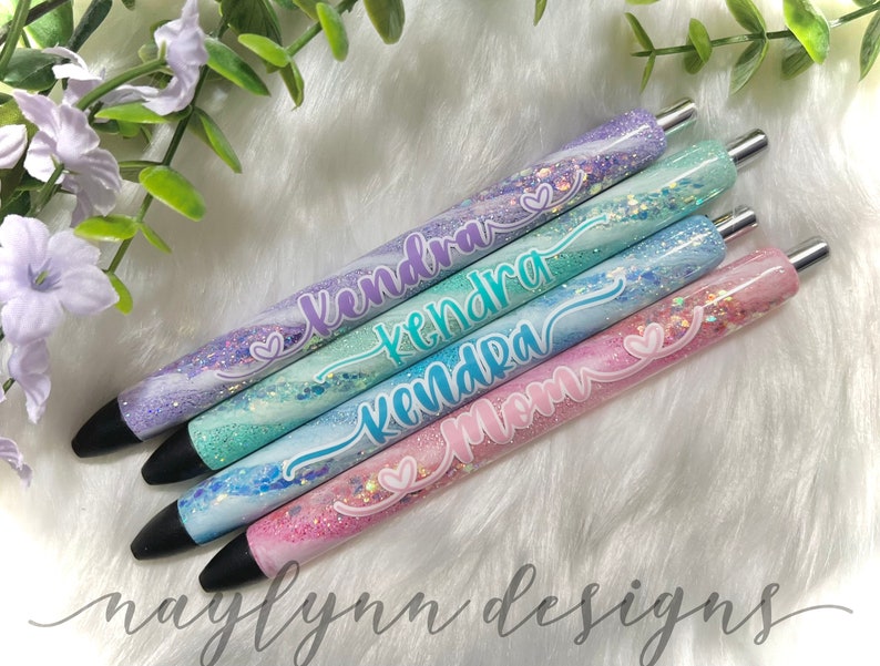 May include: Four personalized pens with glitter and marble designs. The pens are labeled "Kendra", "Mom", and two more "Kendra" names.