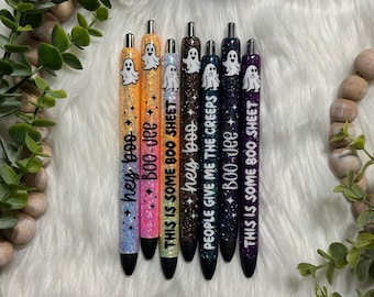 May include: Set of seven glitter pens with ghost and Halloween-themed sayings. The pens are in various colors, including orange, pink, brown, teal, and purple. The sayings on the pens include "hey boo", "Boo-Jee", "THIS IS SOME BOO SHEET", "PEOPLE GIVE ME THE CREEPS", and "THIS IS SOME BOO SHEET".