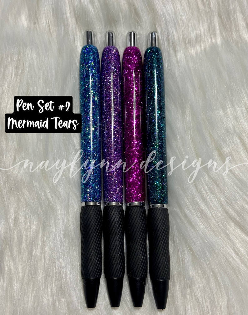 May include: Set of four glitter pens with black ink and silver accents. The pens have a teal, purple, pink, and blue glitter design. The text "Pen Set #2 Mermaid Tears" is visible in the image.