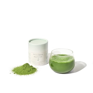 May include: A white cylindrical container with the text "Premium Japanese Matcha" and a green powder next to it. A glass of green matcha tea is in the background.