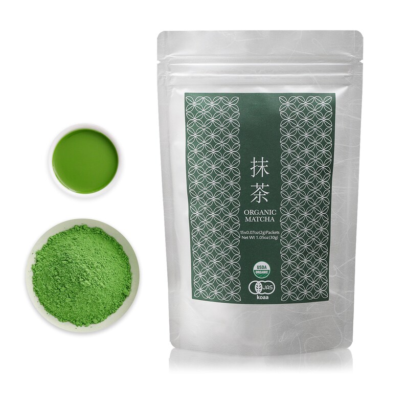 May include: A silver pouch with a green label that says "抹茶 ORGANIC MATCHA" and has a green pattern. The label also includes the text "15x0.07oz(2g) Packets Net Wt 1.05oz(30g)" and has USDA Organic and JAS logos. The pouch is sitting on a white surface. There is a small white bowl with green matcha powder and a small white bowl with green matcha tea.