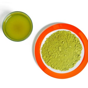 May include: A small glass cup filled with green matcha tea next to a small orange plate with a pile of green matcha powder.