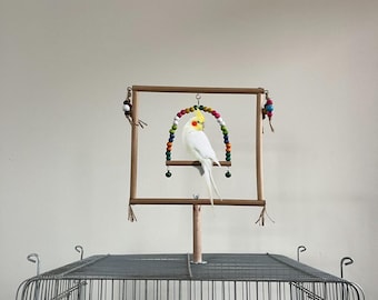 Könnte beinhalten: Ein weißer Nymphensittich sitzt auf einem hölzernen Vogelständer mit bunten Perlenakzenten. Der Ständer ist über einem Vogelkäfig aus Metall aufgehängt. Der Vogel hat einen gelben Kamm und orangefarbene Wangenflecken.