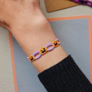 Könnte beinhalten: Ein Halloween-Armband mit orangefarbenen Kürbismotiven, schwarzen Gesichtern und grünen Akzenten, verbunden durch ein hellviolettes gewebtes Band. Das Armband wird am Handgelenk getragen.