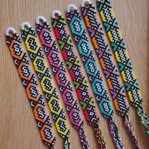 May include: A collection of seven colorful friendship bracelets with intricate geometric patterns. Each bracelet features a unique combination of colors, including orange, pink, green, and blue, against a black background. The bracelets are displayed on a wooden surface.