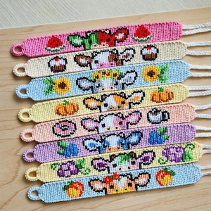May include: A selection of handmade friendship bracelets with vibrant woven patterns. Each bracelet showcases a cow face design alongside various food illustrations, such as watermelon, cupcakes, sunflowers, pumpkins, donuts, blueberries, grapes, and peaches. The bracelets have a loop and a braided string.