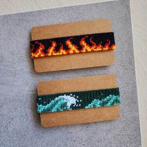 Könnte beinhalten: Zwei Freundschaftsarmbänder auf braunem Karton. Das obere Armband ist schwarz mit orangefarbenen und gelben Flammen. Das untere Armband ist grün mit einem blauen und weißen Wellenmuster.